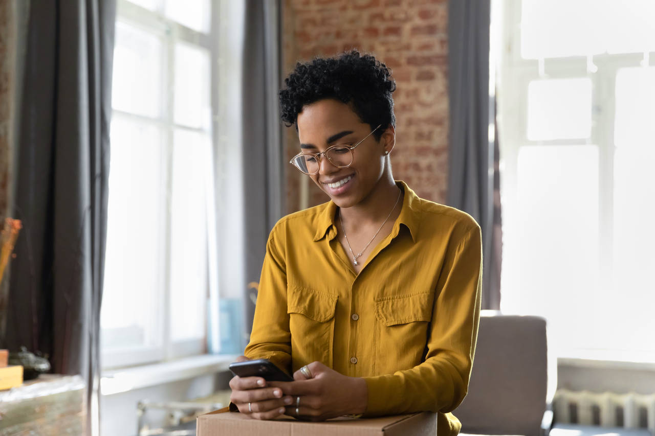 Jeune femme afro-américaine belle et souriante portant des lunettes, laissant un avis positif sur une application mobile d’achat en ligne, s’estimant satisfaite de la commande reçue sur Internet et recommandant le magasin.