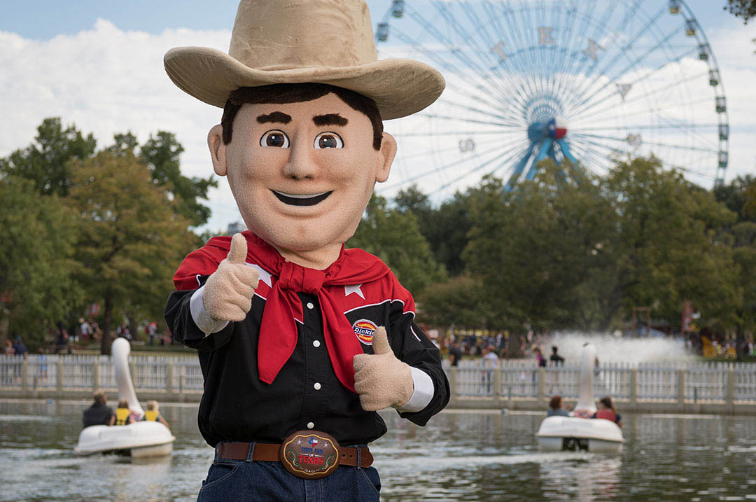 Image of Texas State Faiir cowboy figure thumbs up with ferris wheel and paddle boats in background.